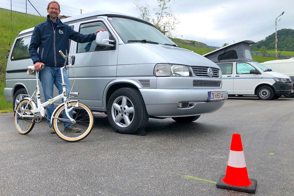 Silberner VW T4 von schräg vorn, der Besitzer steht daneben mit seinem Klappfahhrad von 1970