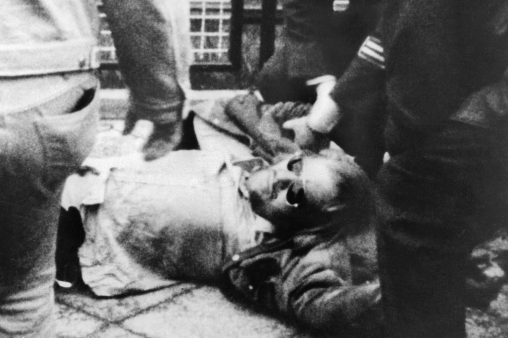 Schwarzweißfoto: Mann mit Sonnenbrille liegt am Boden, Uniformierte halten ihn fest.