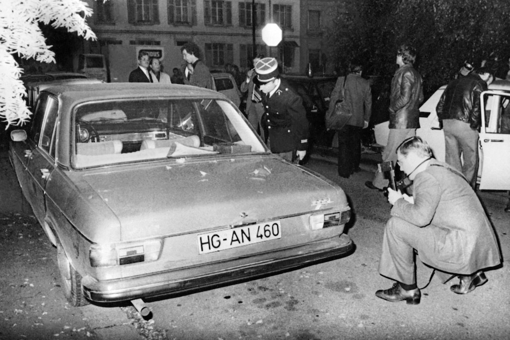 Schwarzweißfoto mit einem Audi 100 (Baureihe C1) von hinten. Ein französischer Polizist in Uniform, ein Fotograf und mehrere Personen sind zu sehen.