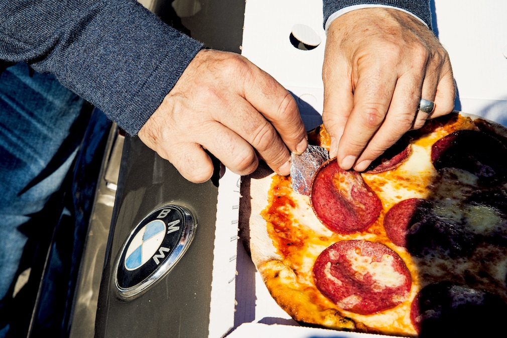 Pizza auf der Kofferraumhaube, eine Hand hält ein Päckchen in Alu-Folie unter einer Salami-Scheibe