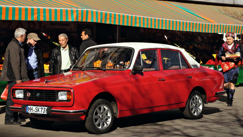 Anadol A2 in Rot von schräg vorn vor einem türkischen Laden in Deutschland, daneben ein Mann in türkischer Tracht