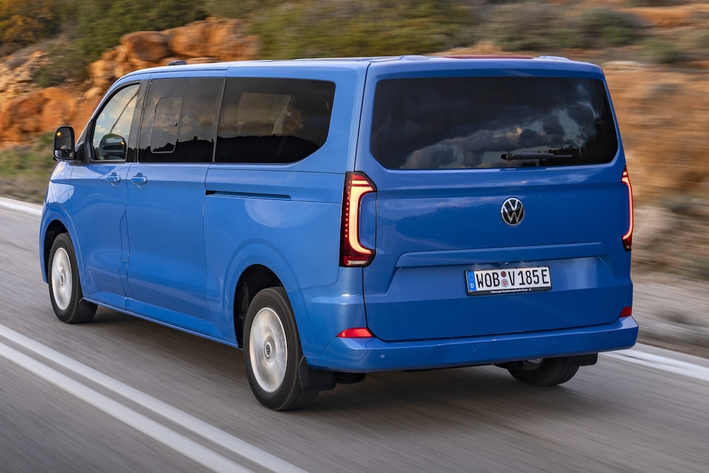 Blauer VW e-Caravelle fährt schräg von hinten über eine Landstraße.