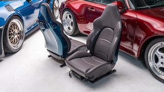Zwei ausgebaute Porsche-Autositze stehen auf dem Boden zwischen zwei Porsche 911