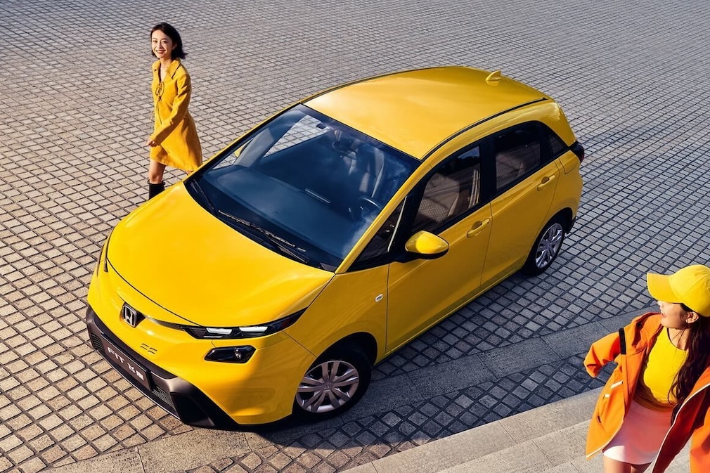 Gelber Honda Fit steht auf einem Platz, zwei Personen gehen daneben.
