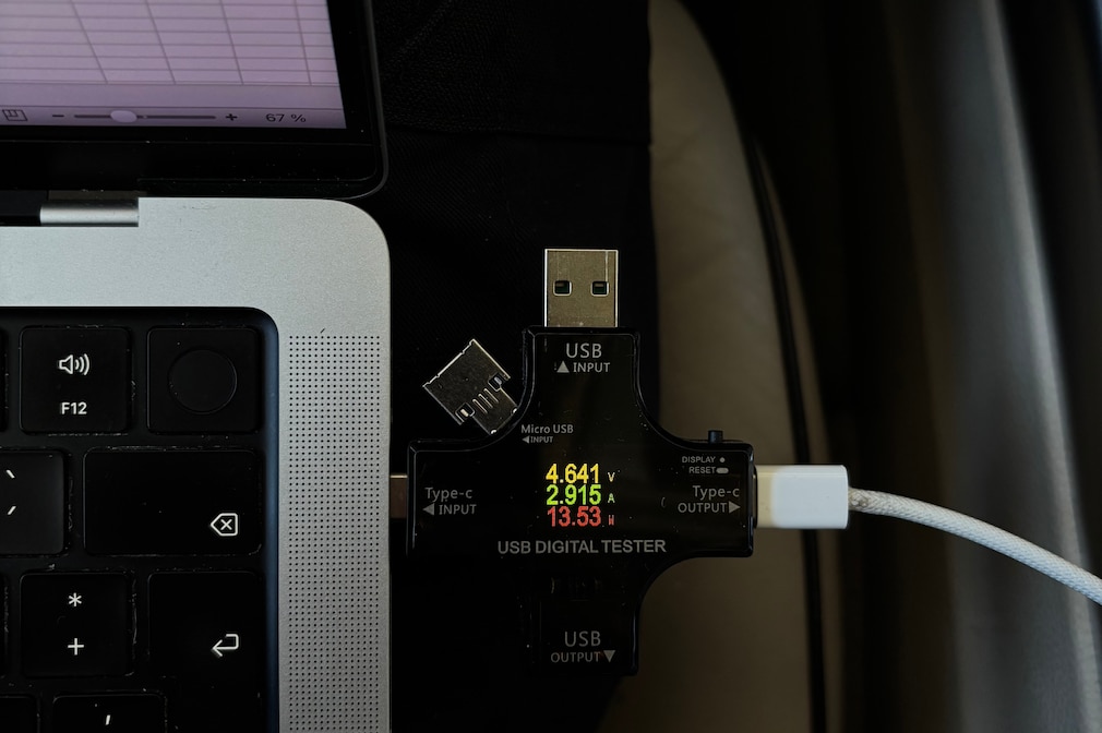 Ein USB-Digitaltester ist zwischen Laptop und Ladekabel angeschlossen und zeigt in Echtzeit Spannung, Stromstärke und Ladeleistung an.
