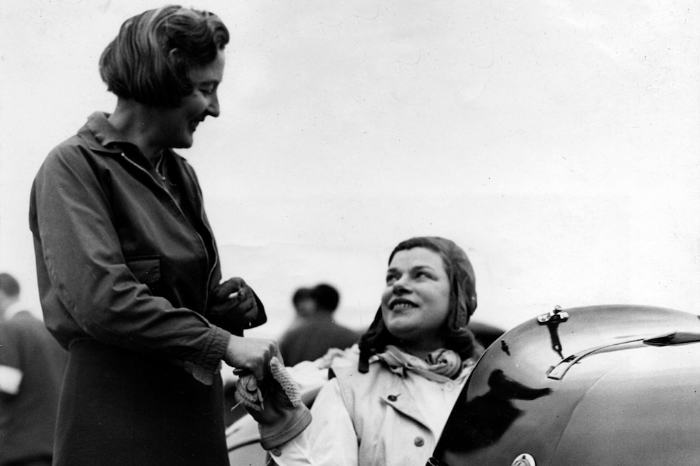 historisches Schwarzweißfoto von zwei Frauen, eine sitzt in einem Rennwagen