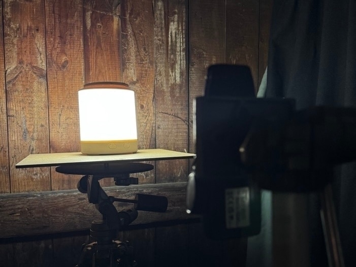 Eine Leuchte steht auf einer Stativfläche vor einer Holzwand. Davor, unscharf, ein Luxmeter als Messinstrument.