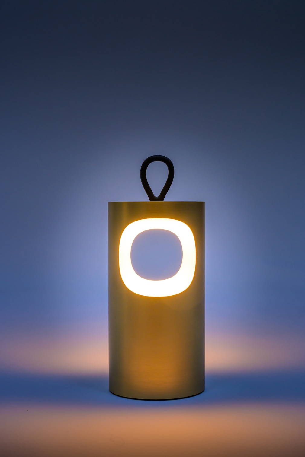Zylinderförmige, minimalistisch gestaltete Leuchte in warmem Gelbton. In ihrer Front befindet sich ein ringförmiges Lichtelement, oben sitzt eine kleine Schlaufe zum Aufhängen. Die Lampe steht vor einem dunkelblauen Hintergrund und strahlt ein sanftes, warmes Licht aus.