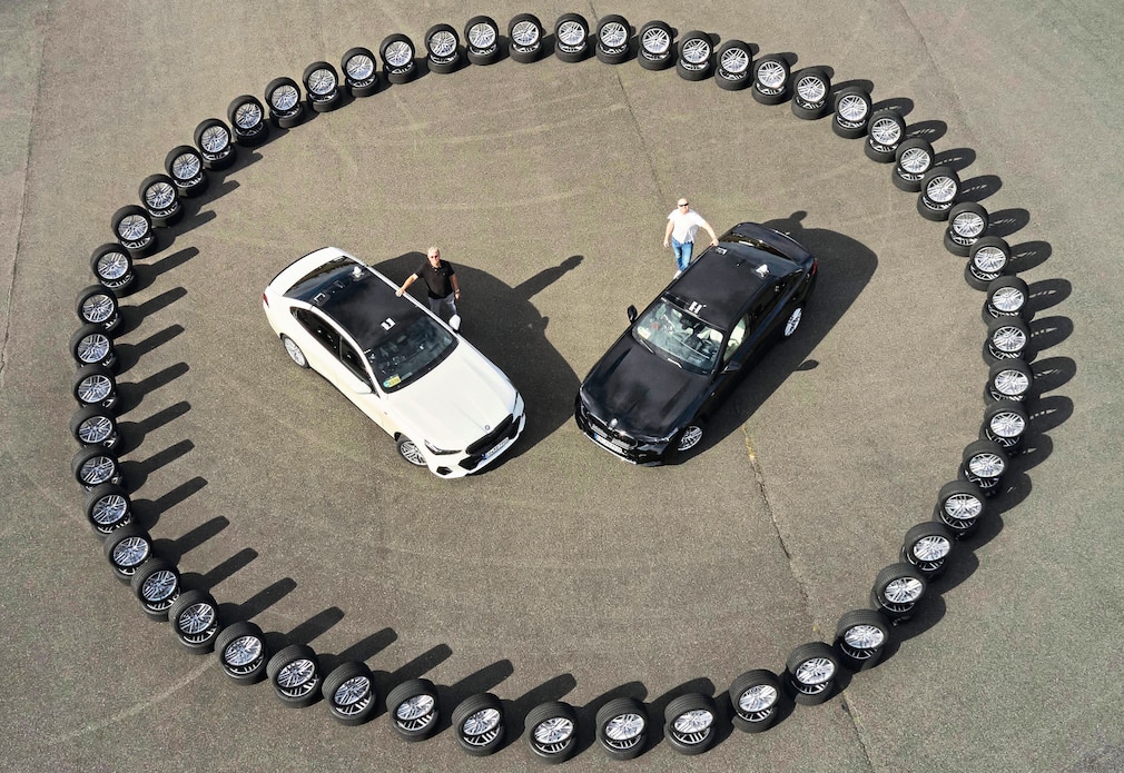 Die AUTO BILD-Reifentester Dierk Möller (links) und Martin Westerhoff stehen neben zwei Testfahrzeugen, die von einem Kreis aus Rädern umgeben sind.