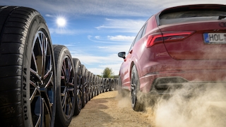 Ein SUV fährt auf losem Untergrund an einer Reihe aneinander gereihter Räder vorbei und wirbelt dabei Staub auf