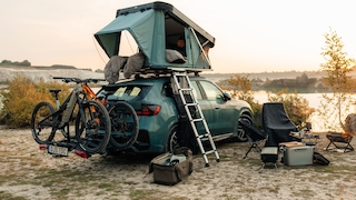 SUV mit Dachzelt, Fahrrädern und Campingausrüstung am Seeufer bei Sonnenuntergang.