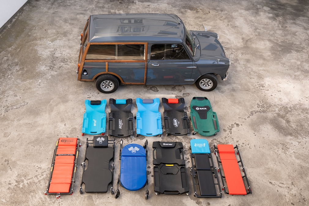 Auf einer Betonfläche stehen mehrere unterschiedliche Rollbretter nebeneinander vor einem klassischen Mini. Die Modelle sind in verschiedenen Bauformen und Farben angeordnet, darunter gepolsterte Liegen, Hartschalenbretter und klappbare Varianten.