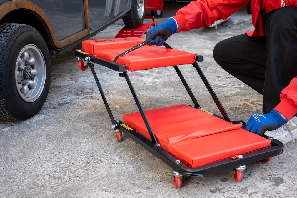 Ein Werkstatt-Rollbrett wird zu einem Hocker umgebaut. Eine Person richtet die klappbare Sitzfläche auf, sodass aus der flachen Liege ein erhöhtes Sitzmöbel mit Rollen entsteht.