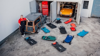 Eine Person lehnt an einem Mini Countryman, der vor einer offenen Garage mit einem Spitfire und einer Werkzeugkiste steht. Im Vordergrund sind 11 verschiedene Werkstatt-Rollbretter zu sehen.