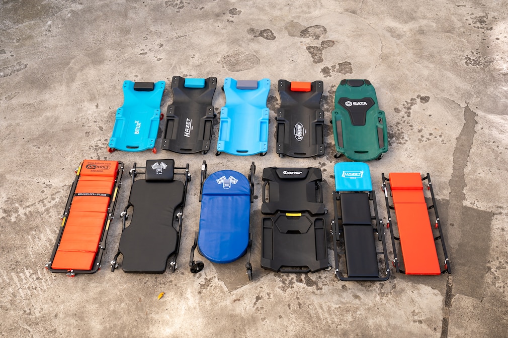 Elf Werkstatt-Rollbretter liegen nebeneinander auf einem Betonboden. Zu sehen sind sowohl Hartschalen- als auch gepolsterte Modelle mit unterschiedlichen Formen, Breiten und Auflagen, die die Unterschiede in Aufbau und Ergonomie zeigen.
