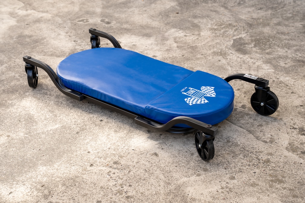 Zu sehen ist ein BGS-Rollbrett mit blau gepolsterter Liegefläche, umlaufendem Stahlrohrrahmen und vier großen, außen montierten Kunststoffrollen auf Betonboden.ahmen montiert, im Hintergrund ist unscharf ein weiterer Teil des Bretts sowie eine behandschuhte Hand zu erkennen.