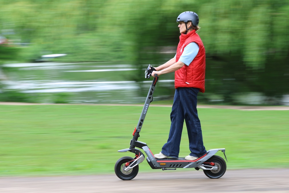 Eine Person fährt mit einem E-Scooter auf einem befestigten Weg in einer Parkanlage.