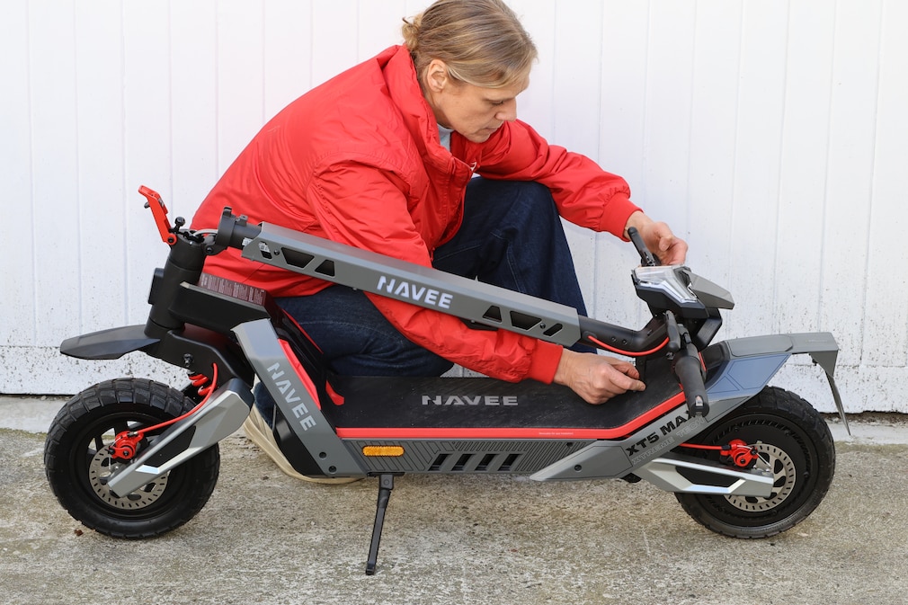 Eine Person kniet neben dem zusammengeklappten E-Scooter und kontrolliert den Klappmechanismus auf sicheren Sitz und Verriegelung.