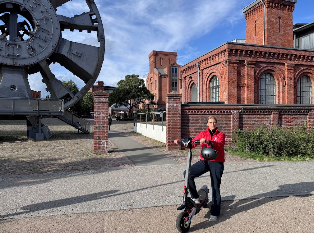 Person steht mit einem Fuß auf dem Trittbrett des Navee XT5 Max. Der Helm hängt am Lenker. Im Hintergrund sind die historischen Backsteingebäude und Industrieanlagen des Museums der Arbeit in Hamburg zu sehen.