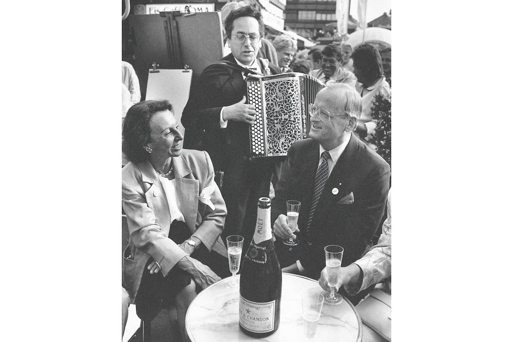 Carl H. Hahn und Ehefrau Marisa Lea Hahn trinken Champagner, hinter ihnen steht ein Musikant mit Akkordeon