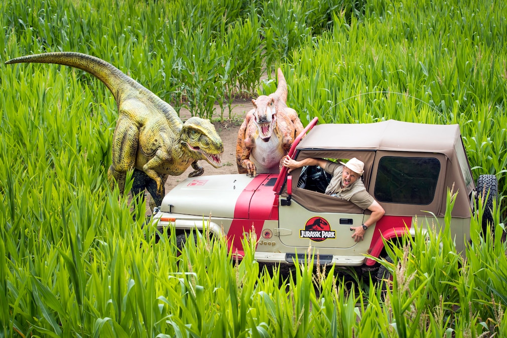 Mann schaut erschrocken aus einem Jeep Wrangler, vor ihm zwei Tyrannosaurus rex
