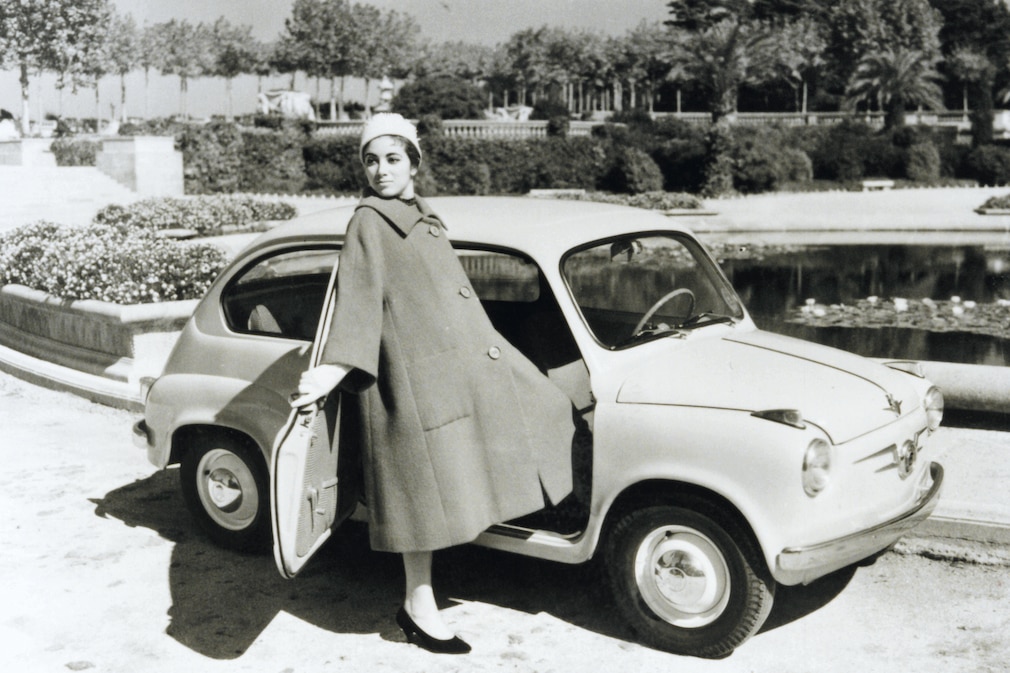Historisches Schwarzweißfoto: Seat 600 mit Frau in langem Mantel an der Beifahrerseite
