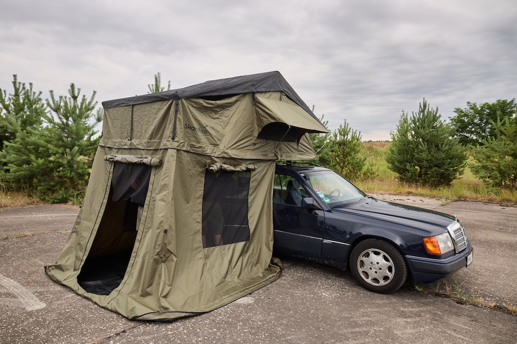 Blauer Meredes W124 mit grünem, auf dem Dach montiertem Dachzelt mit Vorzelt.