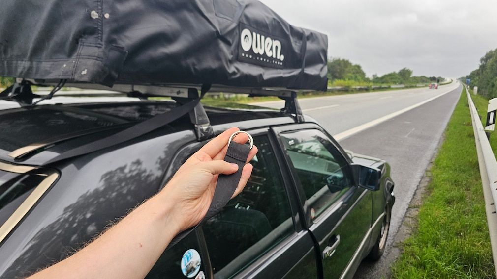 Hand hält gelöste Schnalle eines Dachzelts, Auto steht auf Pannenstreifen der Autobahn
