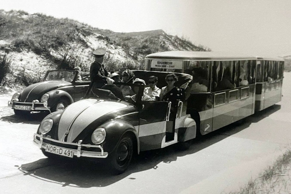 historisches Schwarzweißfoto: VW Käfer Dünen-Express mit zwei Waggons stehend schräg von vorn in den Dünen