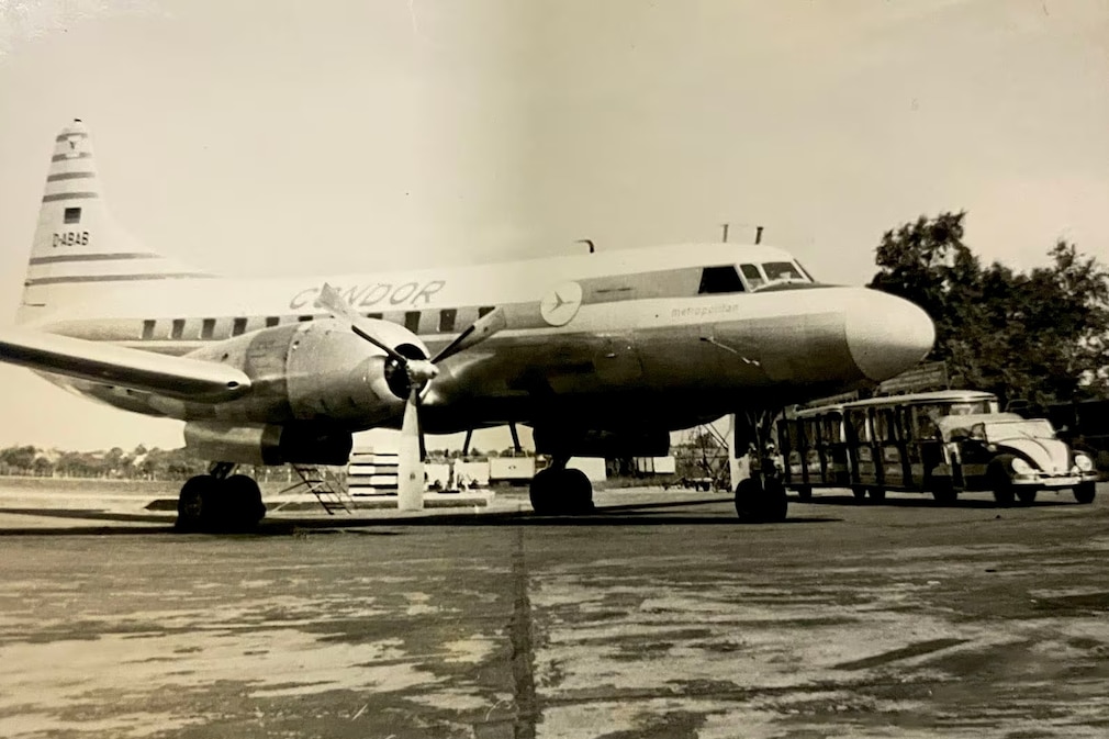 historisches Schwarzweißfoto: VW Käfer Dünen-Express mit zwei Waggons an einem Passagierflugzeug