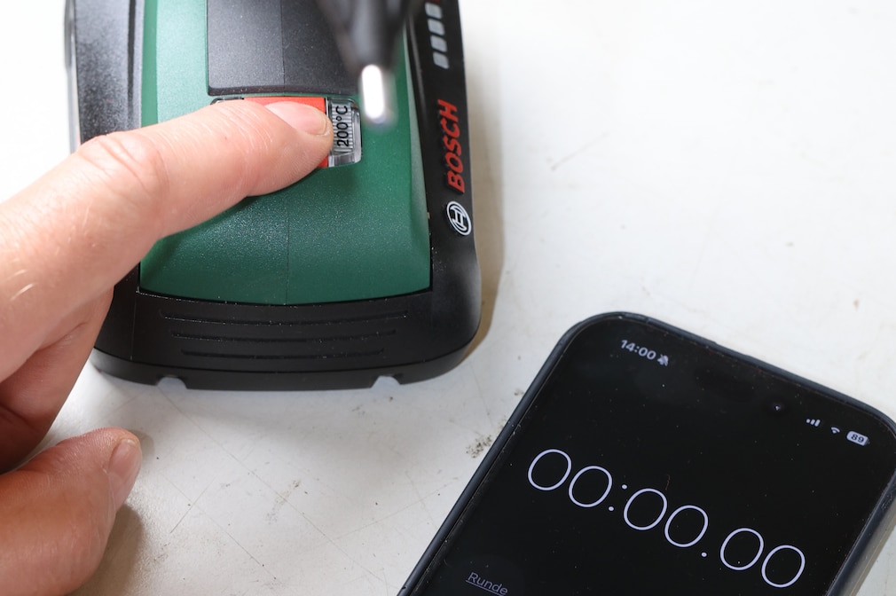 Eine Hand hält ein Smartphone mit Stoppuhr neben eine Heißklebepistole beim Einschaltvorgang zur Zeitmessung der Aufheitzphase.
