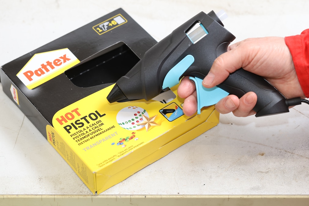 Seitenansicht: Eine Hand hält eine Akku-Heißklebepistole von Pattex über die Verkaufsverpackung mit den Klebestiften.