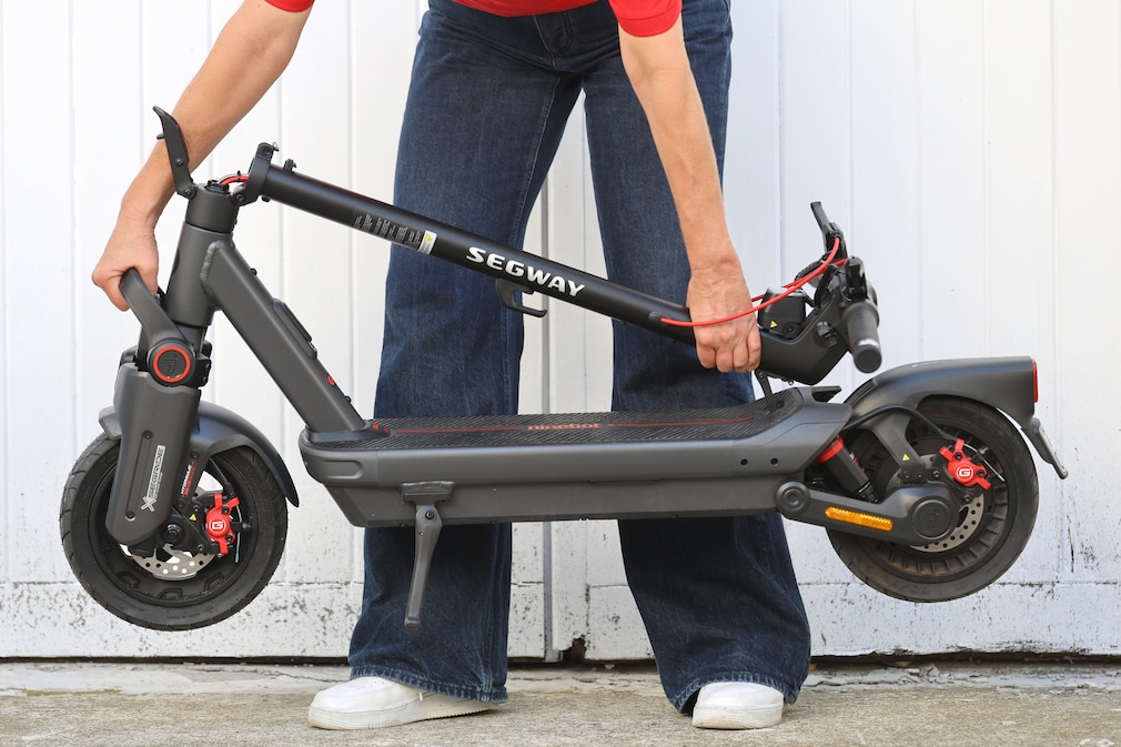 Ausschnitt einer Person die einen E-Scooter im zusammengeklappten Zustand vorne am Griff und hinten am Trittbrett eingehaktem Lenker anhebt