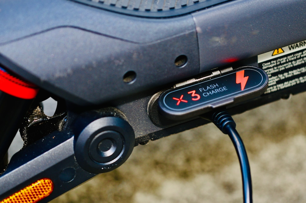Detailaufnahme der Ladebuchse eines E-Scooters mit geöffneter Schutzklappe und angeschlossenem Ladekabel.