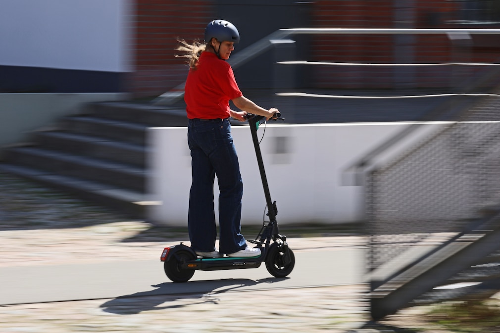 Frau in roter Jacke fährt mit E-Scooter entlang eines Kanals