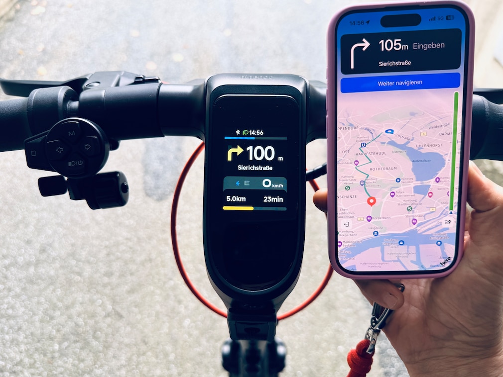 Lenker eines E-Scooters mit Display. Eine Hand hält daneben ein Smartphone. Auf beiden Bildschirmen wird dieselbe Navigationsroute angezeigt.