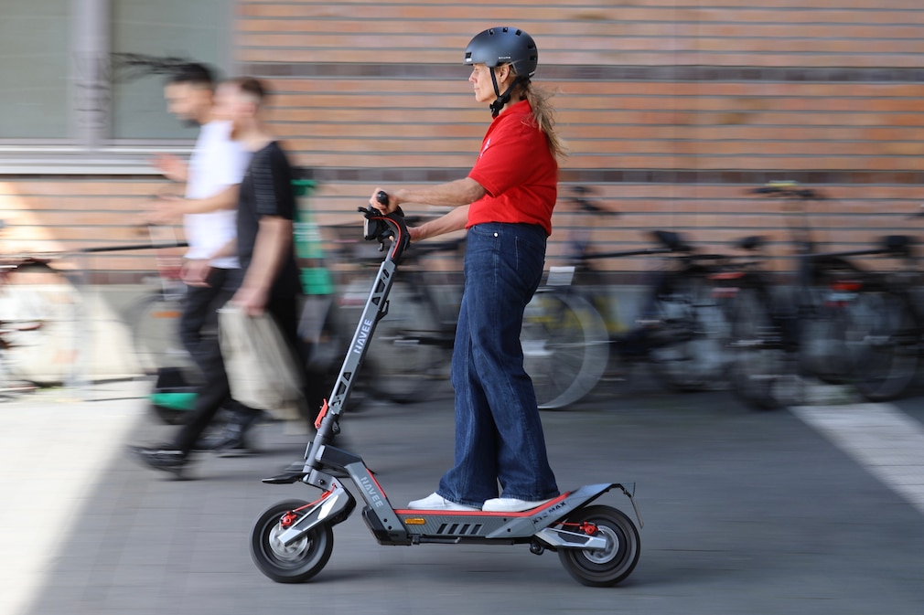 Person fährt mit einem E-Scooter an 2 Personen vorbei im Hintergrund stehen abgestellte Fahrräder vor einer Mauer.