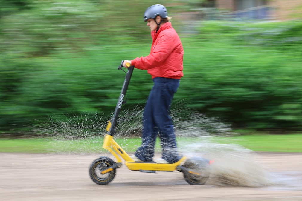 Person fährt mit einem E-Scooter auf unebenem Untergrund durch eine tiefe Pfütze
