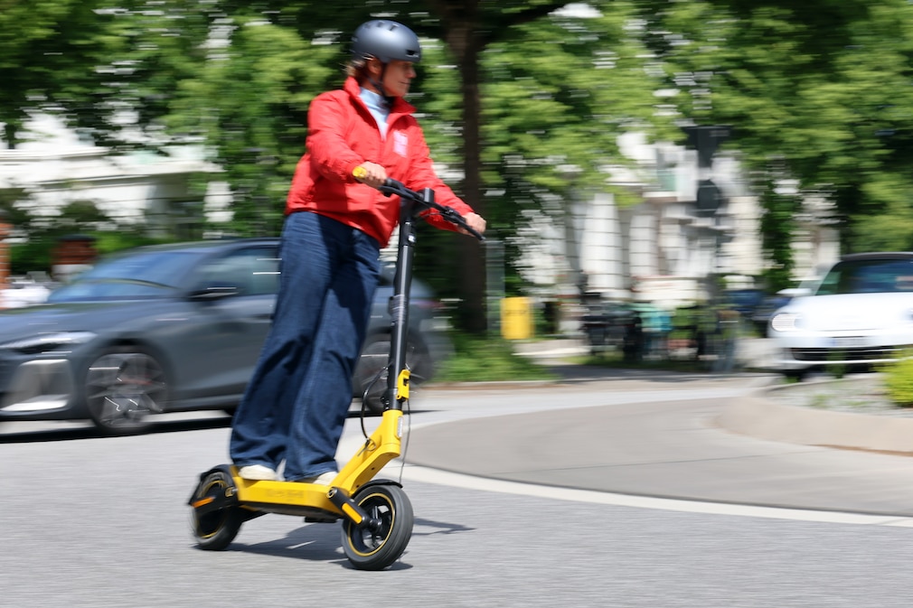 Person fährt mit einem gelben E-Scooter durch eine Kurve auf einer innerstädtischen Straße. Im Hintergrund sind Bäume und Fahrzeuge zu sehen.