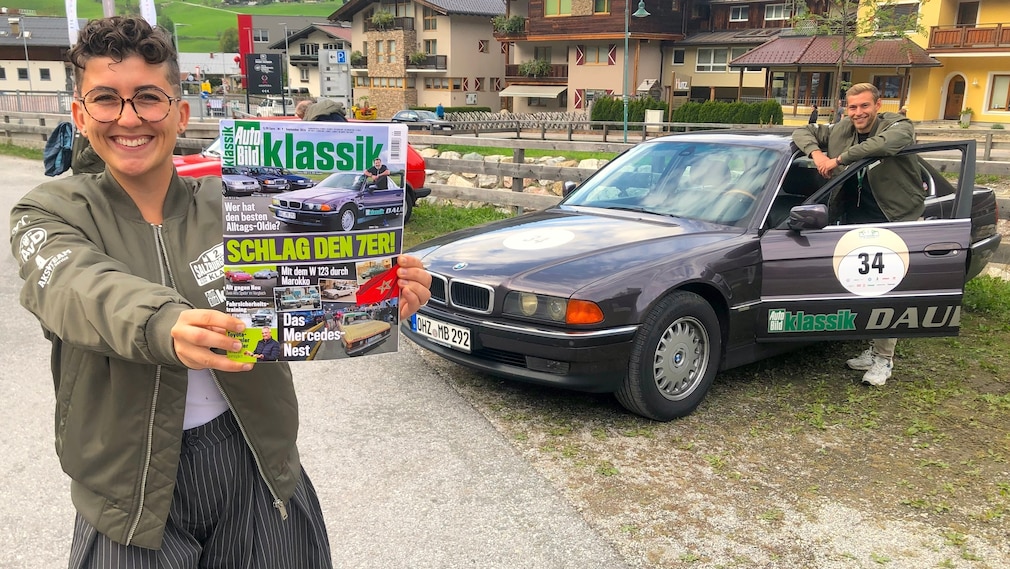 Junge Frau hält eine AUTO BILD KLASSIK mit einem BMW 7er drauf in die Kamera, dahinter steht derselbe BMW in der gleichen Perspektive