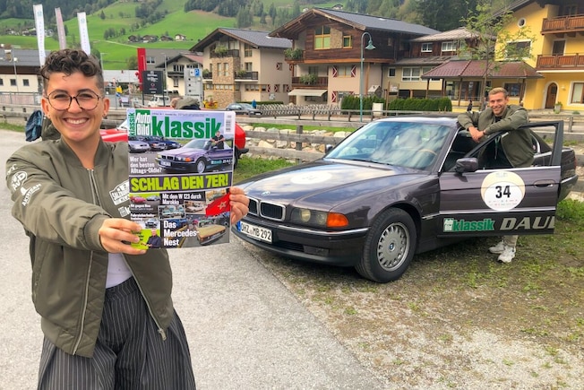 Junge Frau hält eine AUTO BILD KLASSIK mit einem BMW 7er drauf in die Kamera, dahinter steht derselbe BMW in der gleichen Perspektive