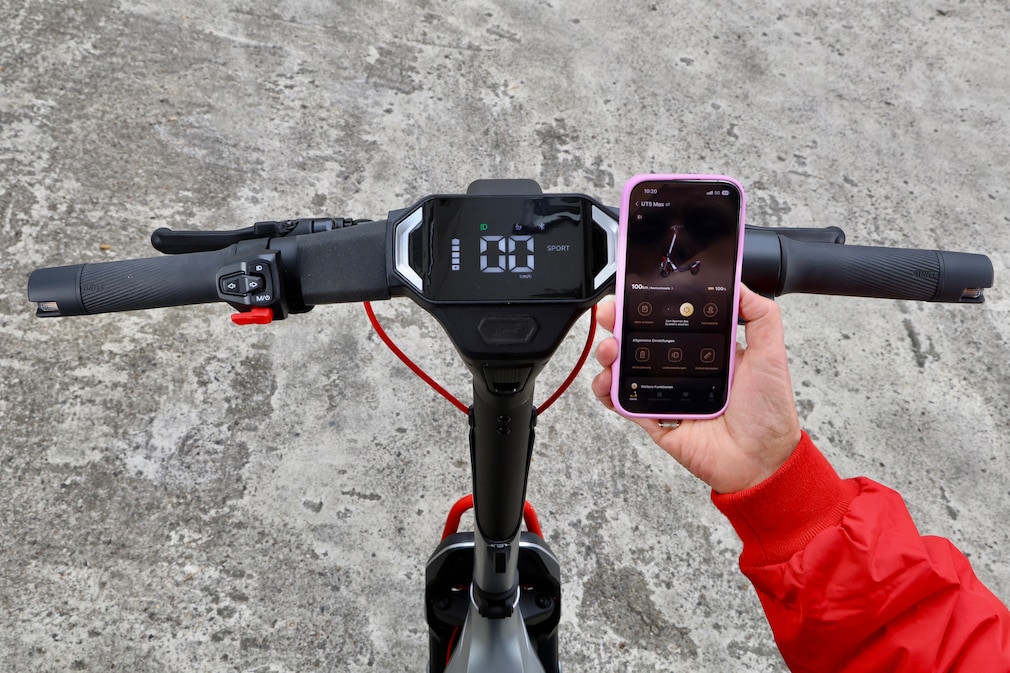 Blick von oben auf den Lenker und das Display eines E‑Scooters. Eine Hand hält daneben ein Smartphone mit geöffneter Fahrzeug-App.