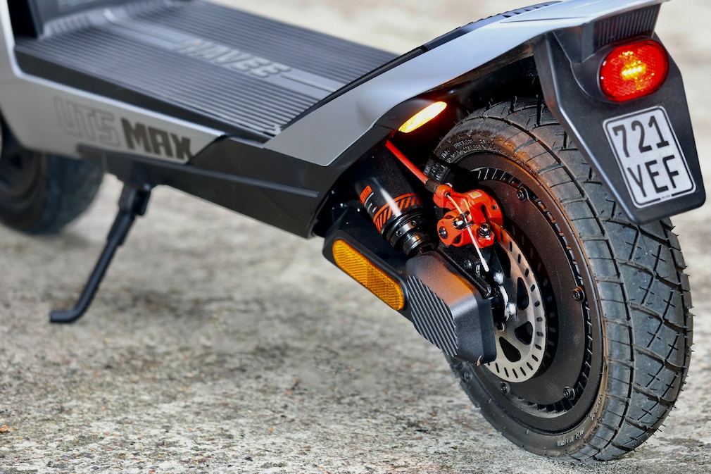 Detailaufnahme des Hinterrads eines E‑Scooters mit Scheibenbremse, Federbein, Rücklicht, Blinker und Reflektor. Das Fahrzeug steht auf seinem Ständer auf einer gepflasterten Fläche.