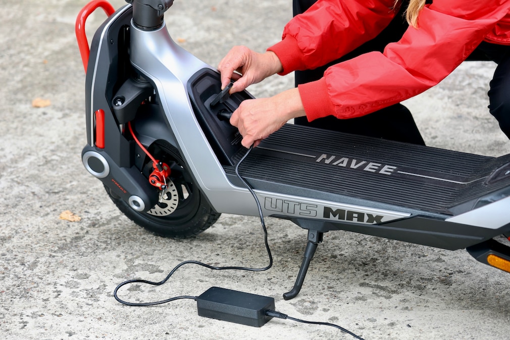 Nahaufnahme eines E-Scooters während des Ladevorgangs. Eine Person in roter Jacke steckt das Ladekabel in die Ladebuchse im Trittbrettbereich. Sichtbar sind das Vorderrad mit Scheibenbremse, ein roter Frontbügel, der ausgeklappte Ständer und das auf dem Boden liegende Netzteil.