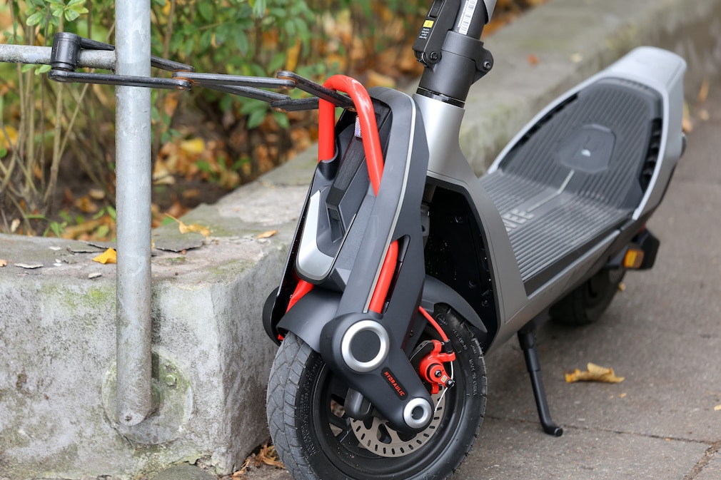 Nahaufnahme eines abgestellten E‑Scooters. Ein schwarzes Faltschloss führt vom roten Frontbügel des Fahrzeugs zu einem Metallpfosten und sichert den Roller. Zu sehen sind außerdem das Vorderrad mit Scheibenbremse, der ausgeklappte Seitenständer, das breite Trittbrett sowie Pflanzen und herbstliches Laub am Fahrbahnrand.
