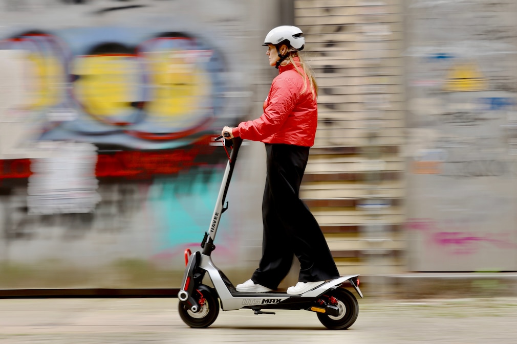 Person mit weißem Helm und roter Jacke fährt auf einem E-Scooter an einer Graffiti-Wand vorbei. Das Fahrzeug ist von der Seite zu sehen, der Hintergrund wirkt durch die Bewegung unscharf.