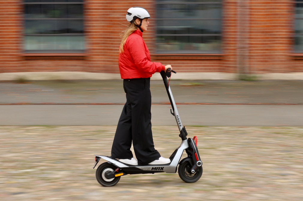 Person mit weißem Helm und roter Jacke fährt auf einem E‑Scooter über Kopfsteinpflaster vor einer Backsteinfassade.