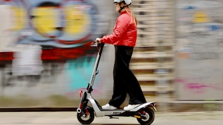 Person mit weißem Helm und roter Jacke fährt auf einem E‑Scooter vor einer unscharfen, mit Graffiti bemalten Wand.