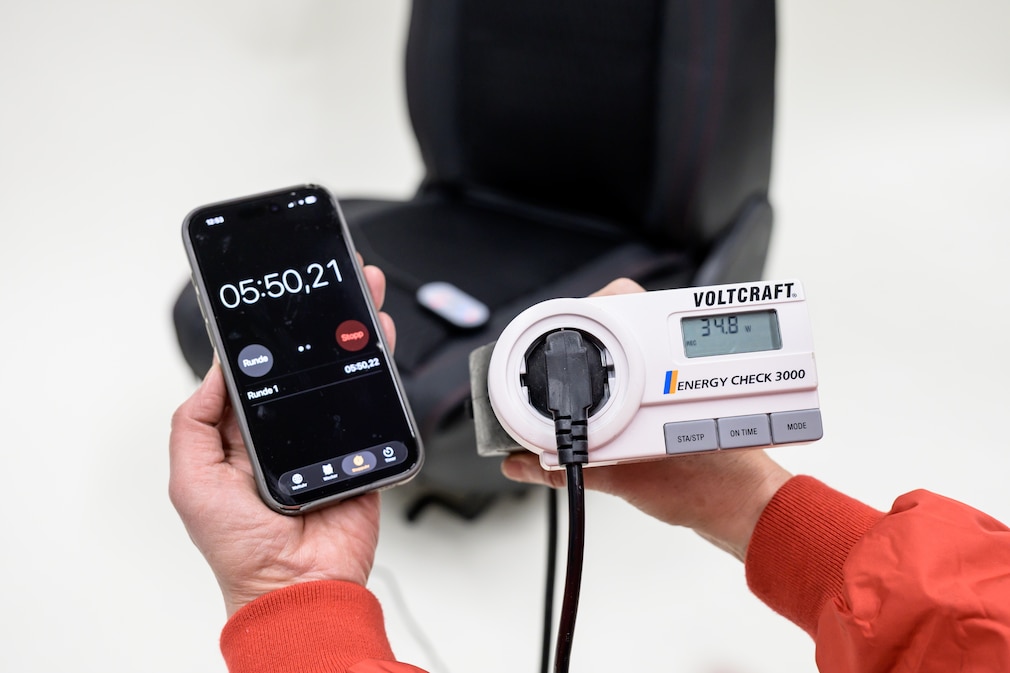 Eine Person hält in einer Hand ein Spannungsmessgerät (Voltmeter) und in der anderen eine Stoppuhr. Vor ihr steht ein ausgebauter Fahrzeugsitz, auf dem eine Sitzheizauflage montiert ist