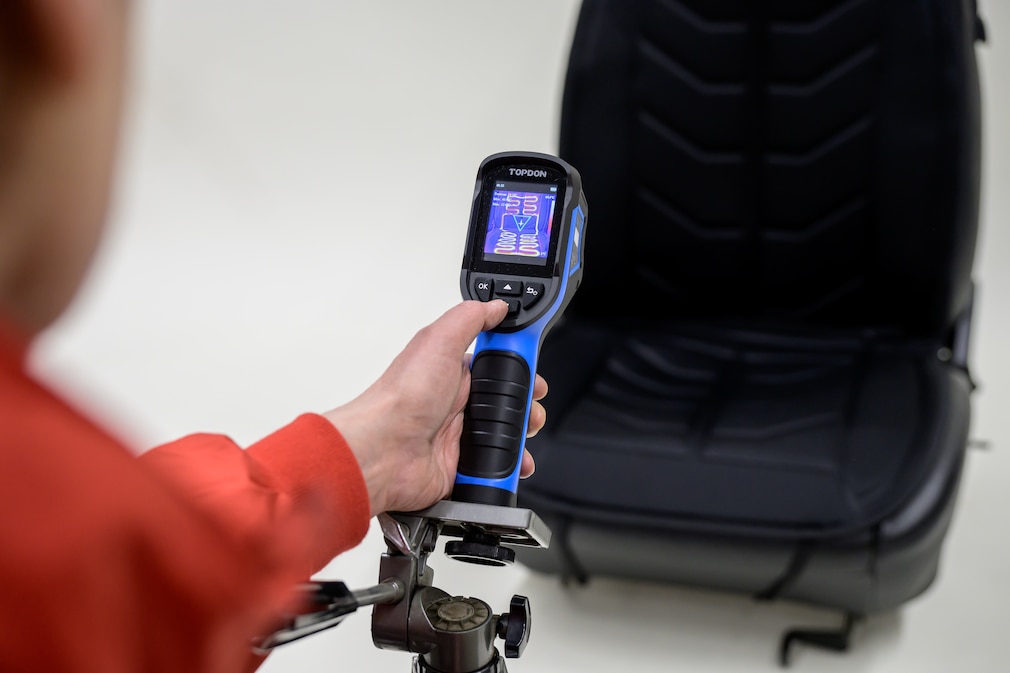 Person richtet eine Wärmebildkamera auf eine Heizmatte die auf einen freistehenden Autositz montiert ist